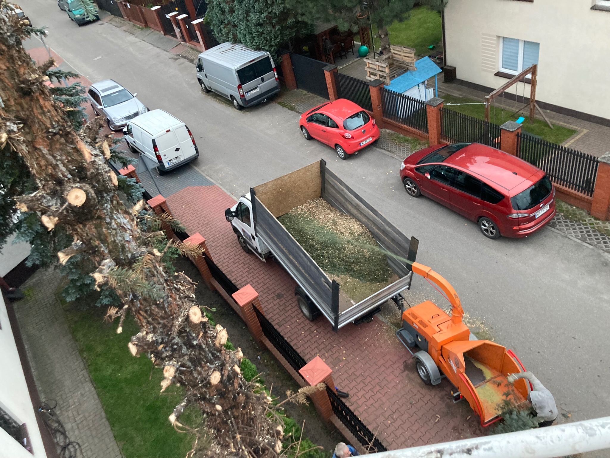 Ciężarówka wypełniona zrębkami drzewnymi obok pomarańczowej rębarki do gałęzi, po wycince drzewa przy ulicy z zaparkowanymi autami. Widok z góry.