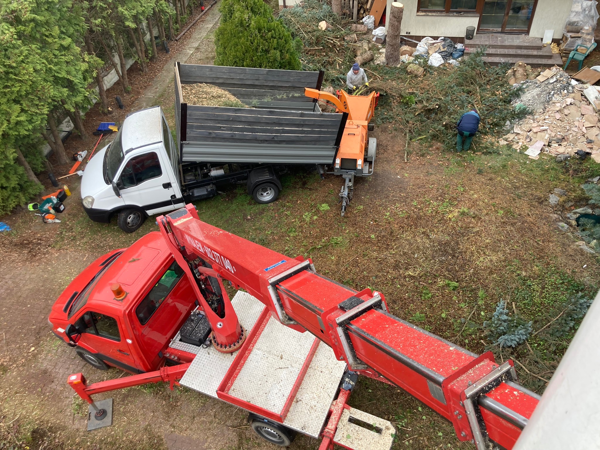 Z góry: czerwony dźwig, biała ciężarówka z przyczepą do rozdrabniania gałęzi, pracownicy porządkujący teren po wycince drzew. Widoczne pnie i gałęzie.