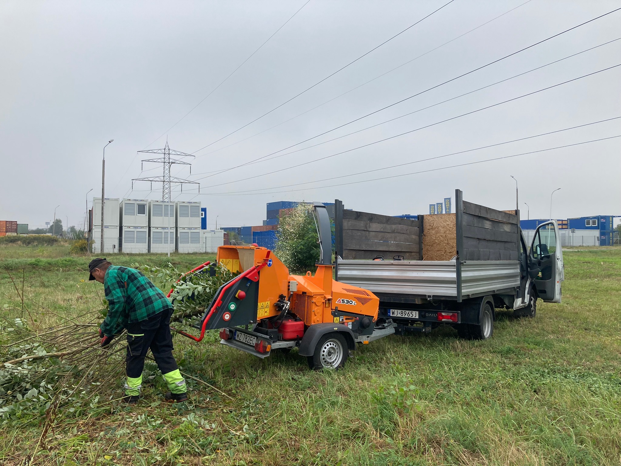 Pracownik w zielonej kracie podaje gałęzie do pomarańczowego rębaka A530L, który jest podpięty do białej ciężarówki na trawiastym terenie. W tle kontenery i słupy wysokiego napięcia.