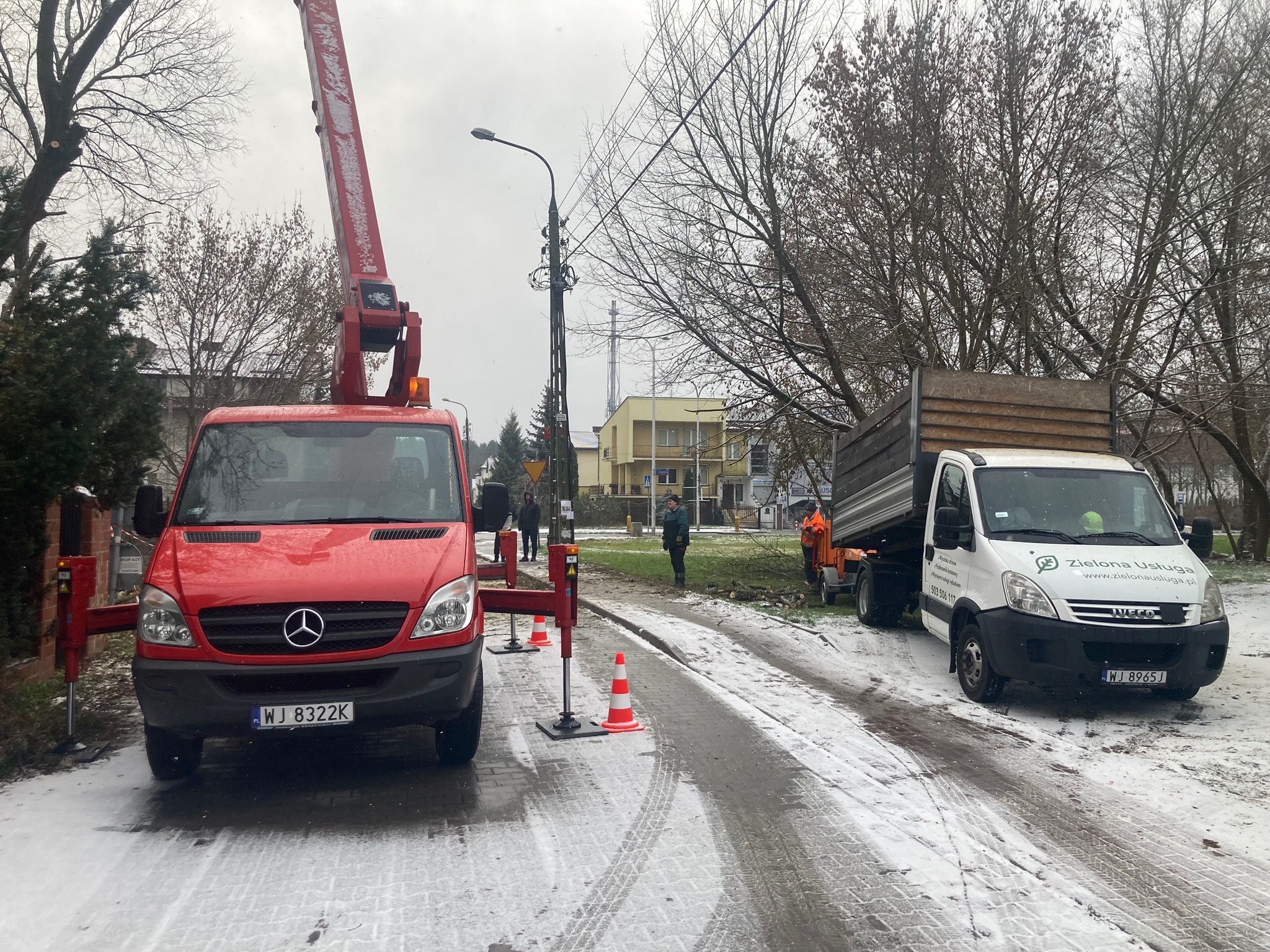 Czerwony podnośnik koszowy Mercedes i biała ciężarówka Iveco na ośnieżonej ulicy podczas prac związanych z pielęgnacją drzew. Widoczni pracownicy w odblaskowych ubraniach.