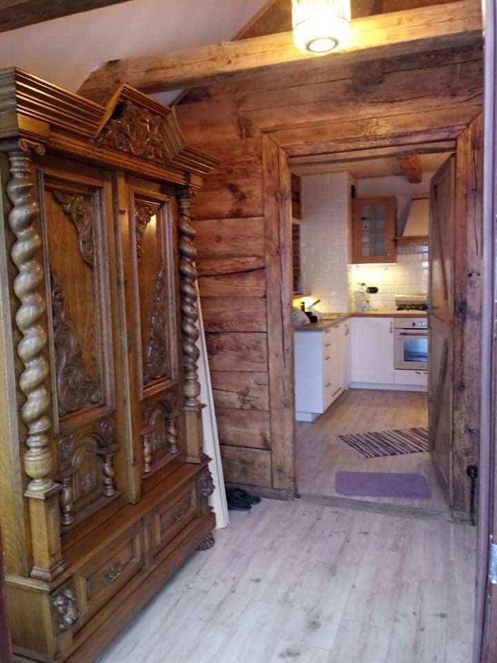 Wnętrze domu z drewnianymi belkami stropowymi i ozdobną szafą, widok na nowoczesną kuchnię z białymi szafkami i płytkami.