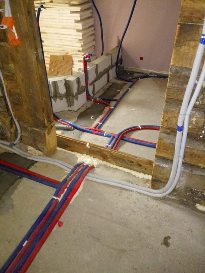 Instalacja wodno-kanalizacyjna w trakcie montażu: czerwono-niebieskie rury prowadzone wzdłuż drewnianej konstrukcji i betonowej podłogi, z widocznymi bloczkami betonowymi i pianką montażową.