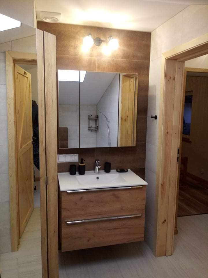 Nowoczesna łazienka z drewnianymi akcentami: szafka pod umywalką z dwoma szufladami, lustro w szafce z oświetleniem nad nim, brązowe płytki na ścianie, widoczny fragment kabiny prysznicowej...