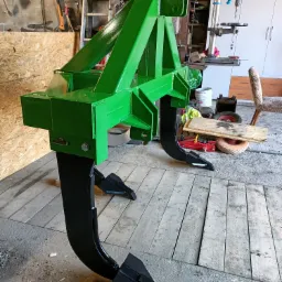 Świeżo pomalowany, zielony kultywator z czarnymi zębami, wykonany przez spawacza z Nakła nad Notecią, stoi w warsztacie na drewnianej podłodze.