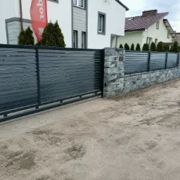 Nowoczesna brama wjazdowa przesuwna, z szarych paneli poziomych, osadzona na słupkach z szarej kostki, na tle domu z czerwonym logo firmy.