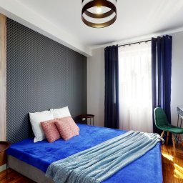 Wnętrze Pod Klucz - Nowoczesna sypialnia z łóżkiem w niebieskiej tonacji, szarą ścianą z geometrycznym wzorem, dużym lustrem z drewnianą ramą i okrągłą lampą sufitową.