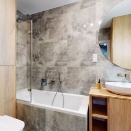 Wnętrze Pod Klucz - Nowoczesna łazienka z wanną, kabiną prysznicową, umywalką nablatową na drewnianej szafce, okrągłym lustrem i szarymi płytkami imitującymi beton.