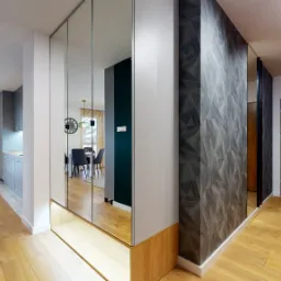 Nowoczesny przedpokój z lustrzaną szafą wnękową, podświetlaną półką, panelami ściennymi z geometrycznym wzorem i drewnianymi drzwiami wejściowymi.