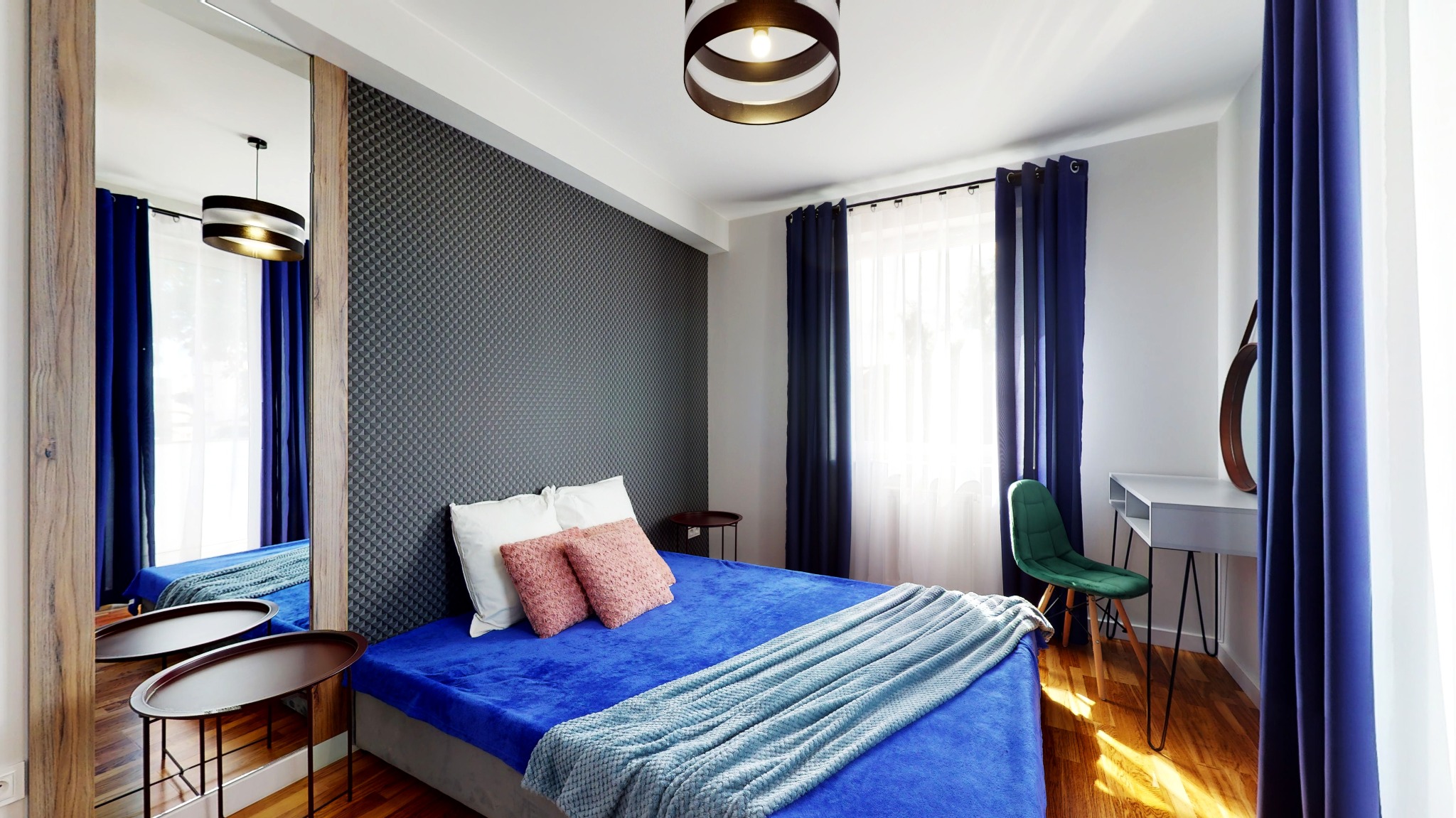 Nowoczesna sypialnia z łóżkiem w niebieskiej tonacji, szarą ścianą z geometrycznym wzorem, dużym lustrem z drewnianą ramą i okrągłą lampą sufitową.