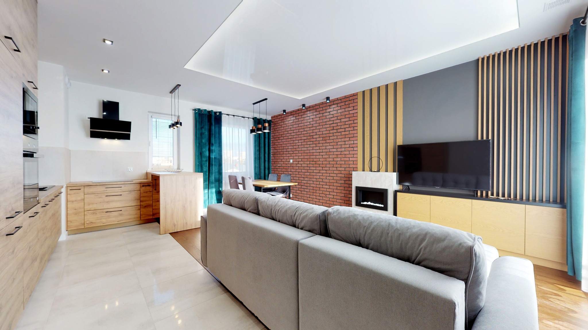 Nowoczesne wnętrze salonu z aneksem kuchennym w Kielcach, z ceglaną ścianą, drewnianymi lamelami i szarym narożnikiem, oświetlone punktowymi lampami.