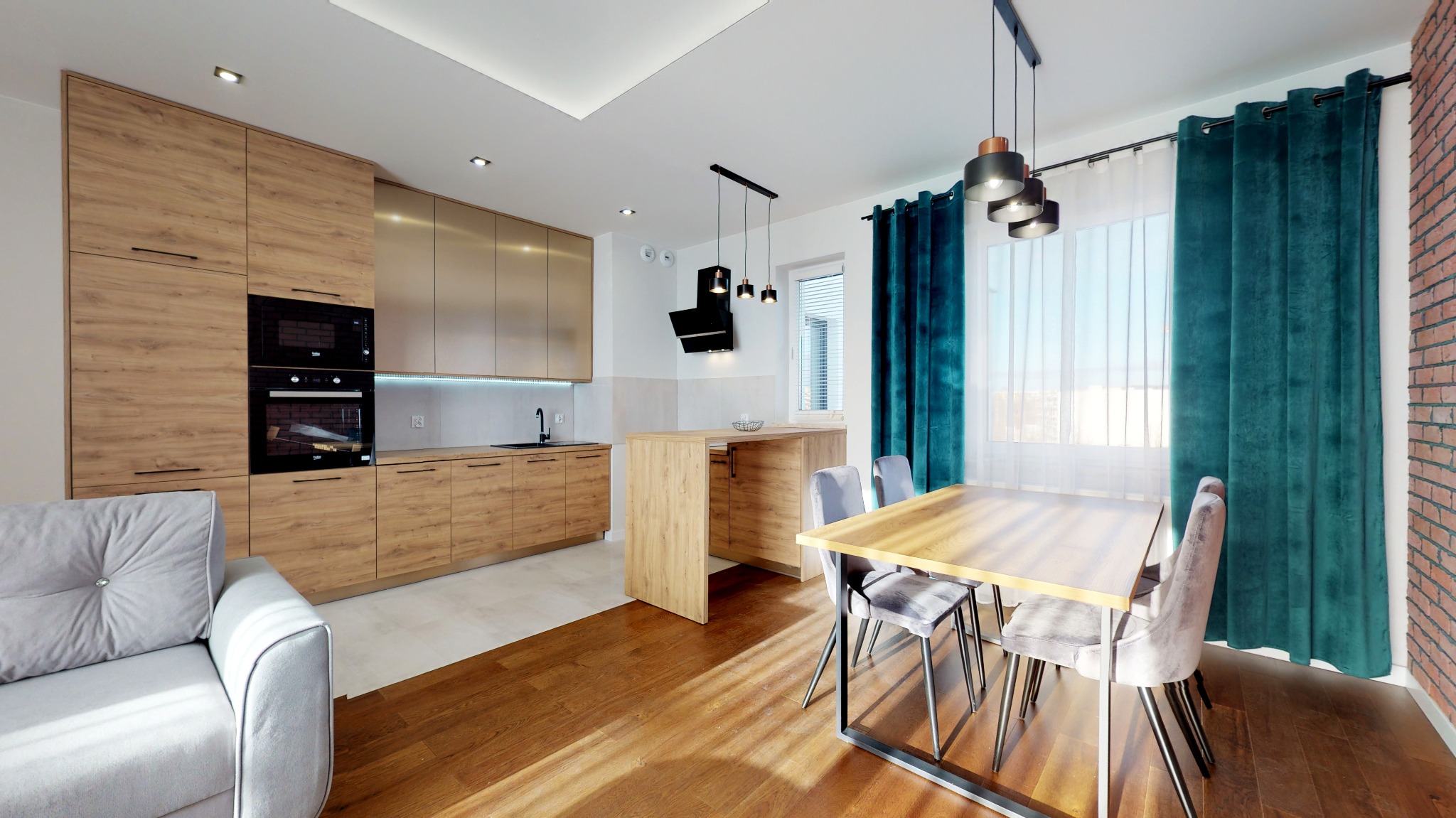 Jasna kuchnia z drewnianymi szafkami, wyspą i stołem jadalnym, akcentowana turkusowymi zasłonami i ceglaną ścianą