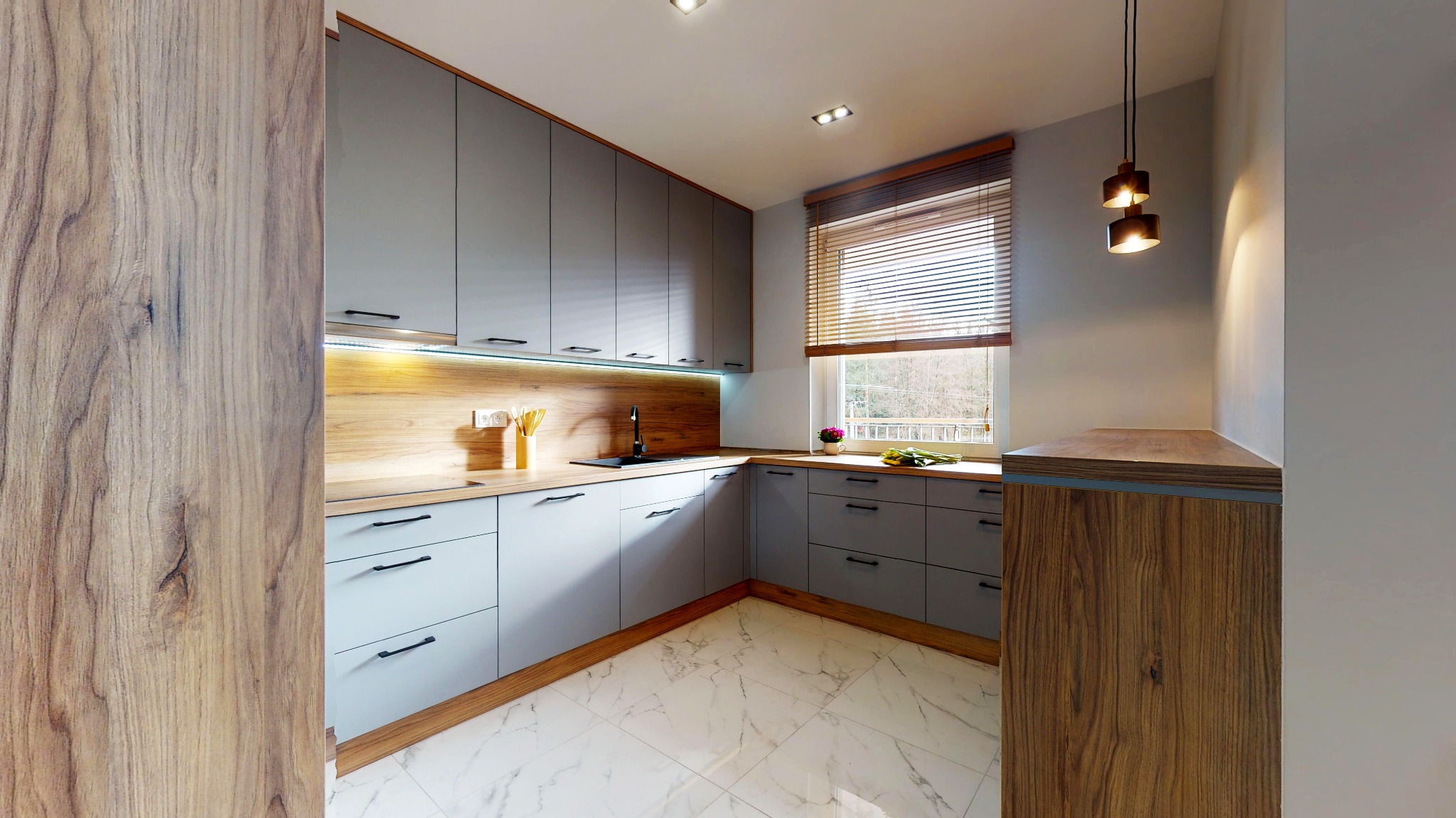 Nowoczesna kuchnia z szarymi szafkami, drewnianym blatem i marmurową podłogą, oświetlona punktowymi lampami sufitowymi i lampą wiszącą, z widokiem na zewnątrz przez okno z drewnianymi żaluzjami.