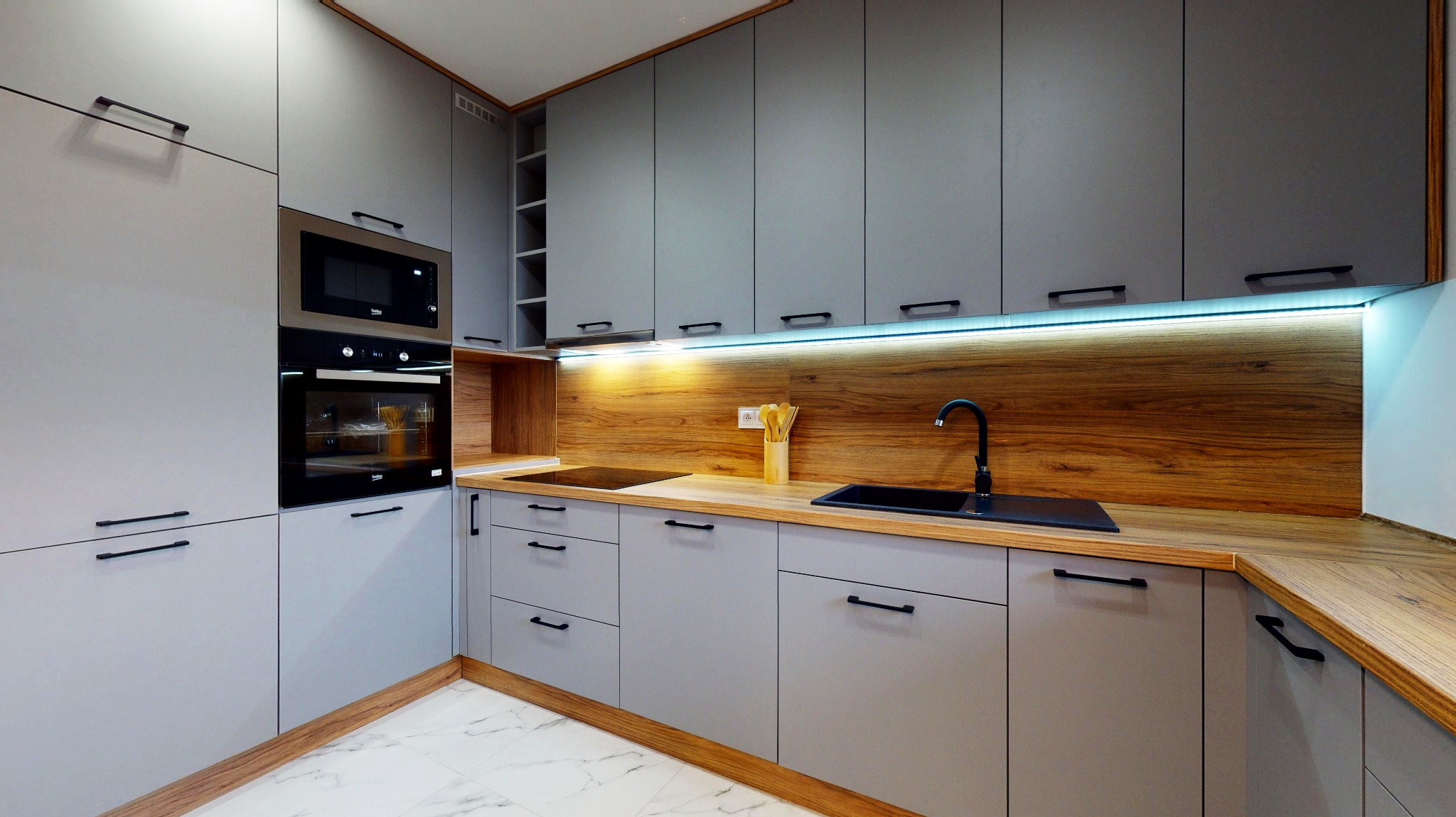 Szara, nowoczesna kuchnia z drewnianymi akcentami: szafki z czarnymi uchwytami, drewniany blat i panel ścienny, oświetlenie LED pod szafkami górnymi, marmurowa podłoga.