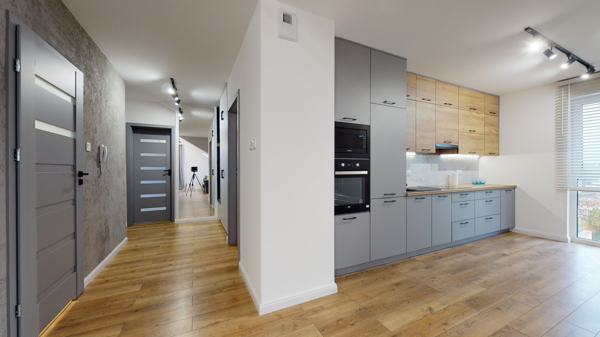 Nowoczesne wnętrze mieszkania z szarymi drzwiami i kuchnią w odcieniach szarości i drewna, parkiet na podłodze, widok na korytarz z lustrem i statywem w tle.