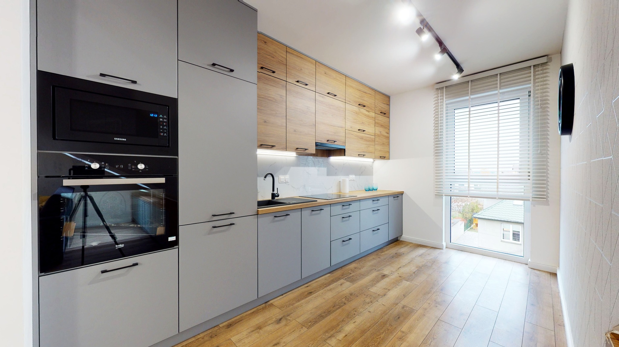 Nowoczesna kuchnia z szarymi szafkami dolnymi i drewnianymi górnymi, czarnym zlewem i armaturą, jasną podłogą oraz oknem z żaluzjami.