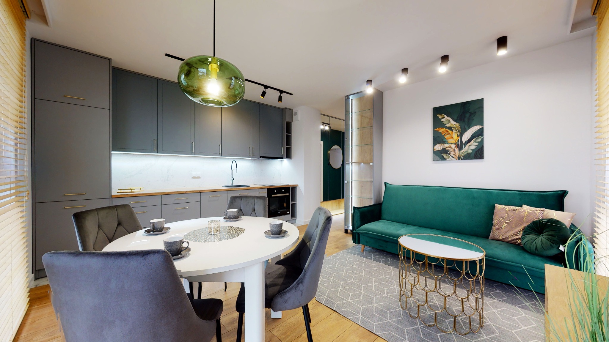 Nowoczesne wnętrze: szare szafki kuchenne z drewnianym blatem, okrągły biały stół z szarymi krzesłami, zielona sofa z dekoracyjnymi poduszkami, złoty stolik kawowy i szary dywan w geometryczne wzory.