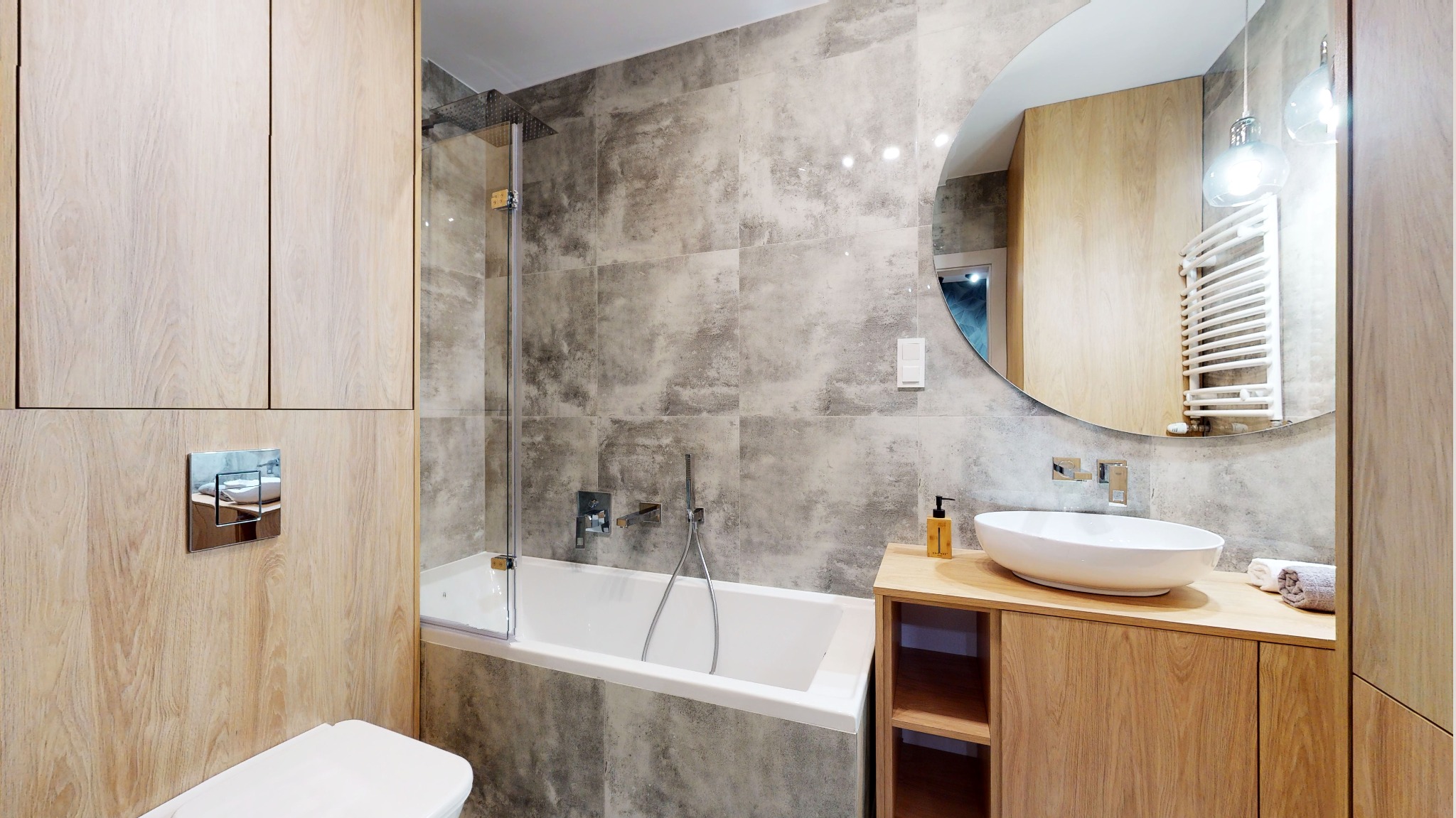 Nowoczesna łazienka z wanną, kabiną prysznicową, umywalką nablatową na drewnianej szafce, okrągłym lustrem i szarymi płytkami imitującymi beton.