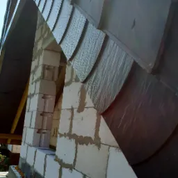 Wznoszenie komina z bloczków betonu komórkowego przy nowym dachu z łupka bitumicznego, widoczne odeskowanie i fragment więźby dachowej.