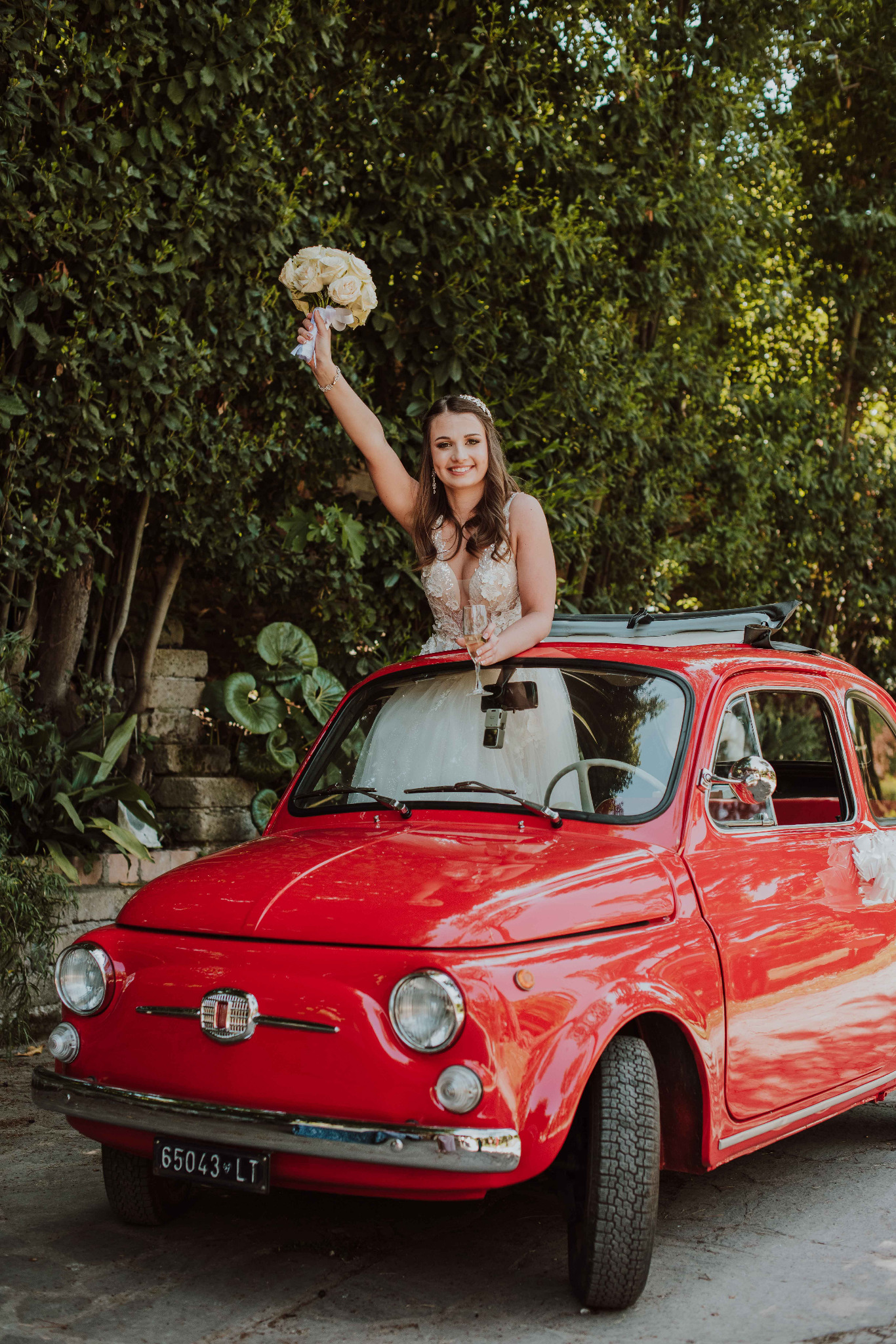 Uśmiechnięta panna młoda w koronkowej sukni, stojąca w otwartym dachu czerwonego Fiata 500, trzymająca bukiet białych róż i kieliszek szampana na tle zieleni.