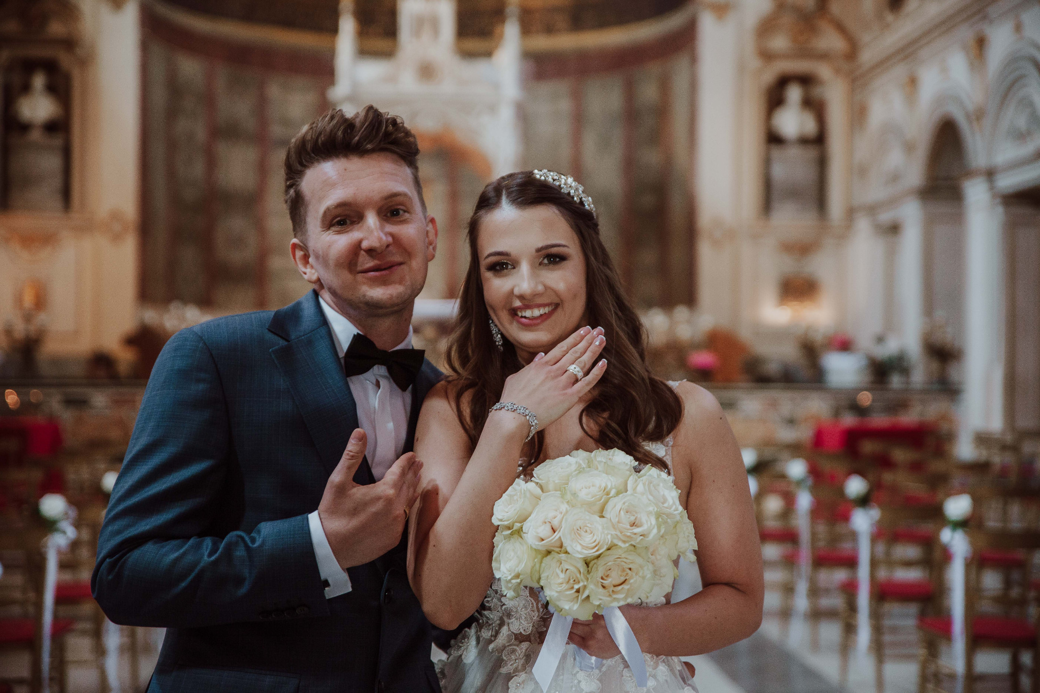 Uśmiechnięta para młoda w kościele, panna młoda trzyma bukiet białych róż i prezentuje pierścionek, pan młody w granatowym garniturze z muchą.