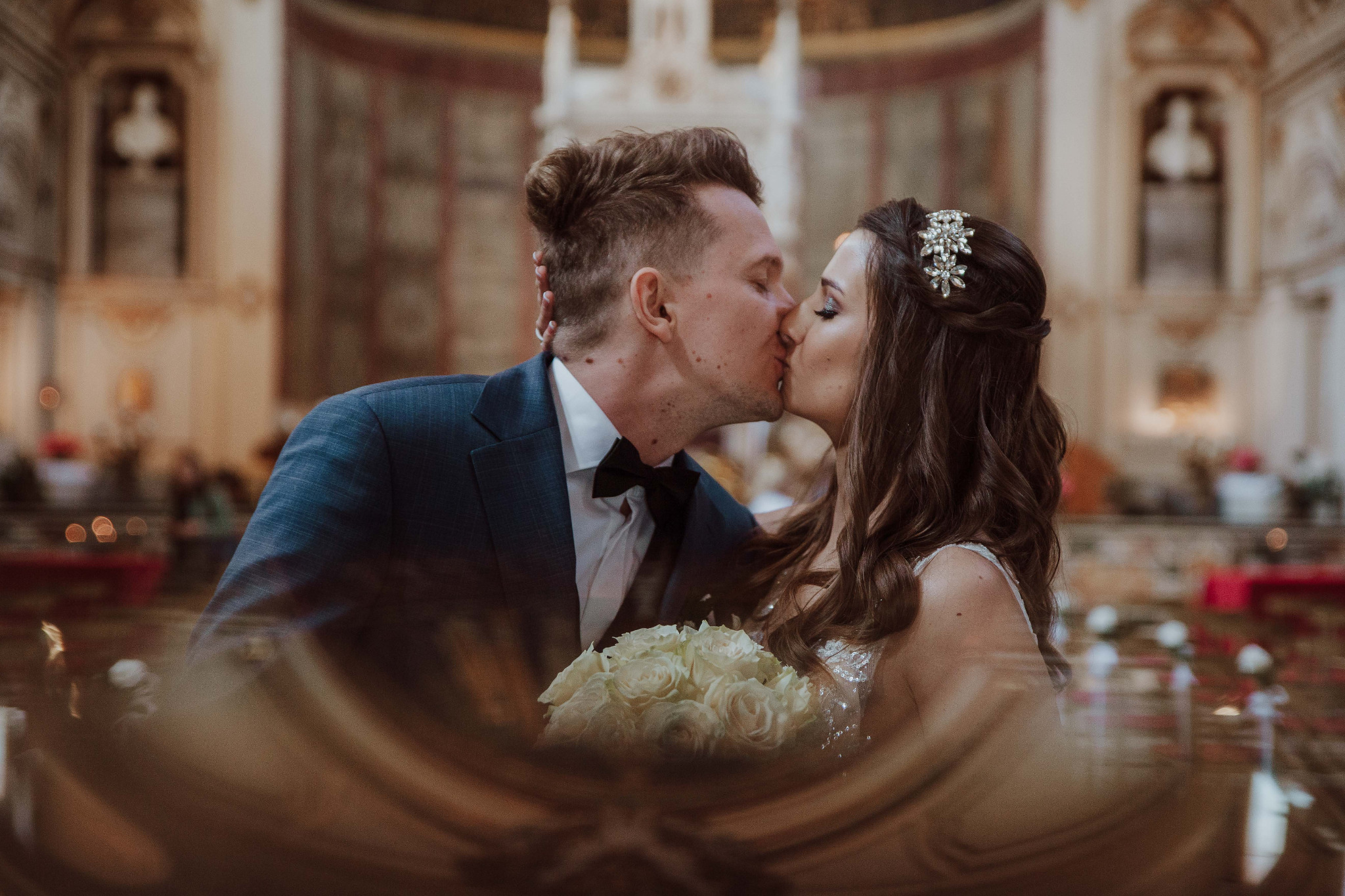 Pocałunek nowożeńców w kościele, panna młoda z bukietem białych róż i ozdobą we włosach, pan młody w granatowym garniturze i muszce.