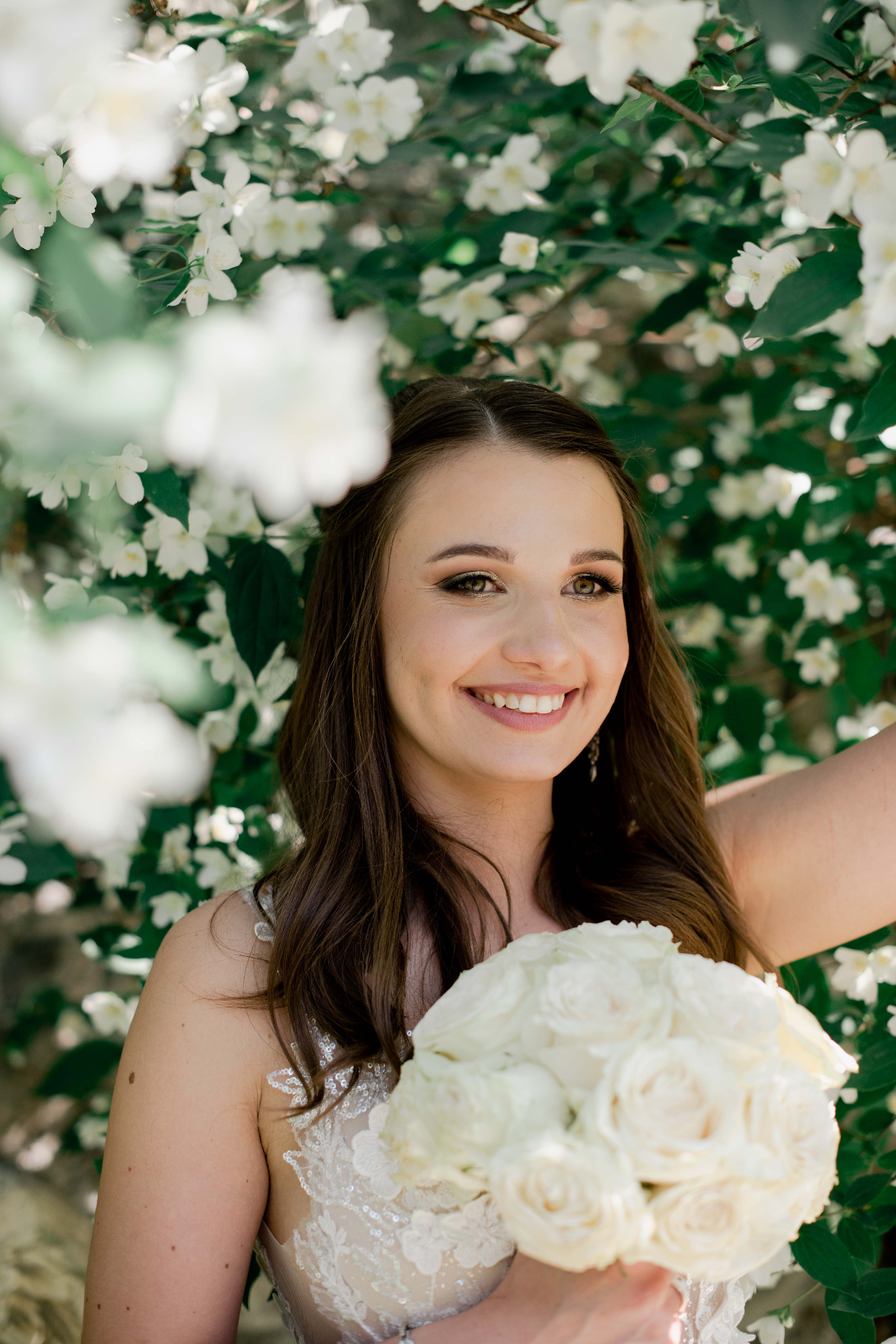 Uśmiechnięta panna młoda z bukietem białych róż na tle kwitnącego jaśminu, delikatny makijaż podkreślony złotym cieniem.