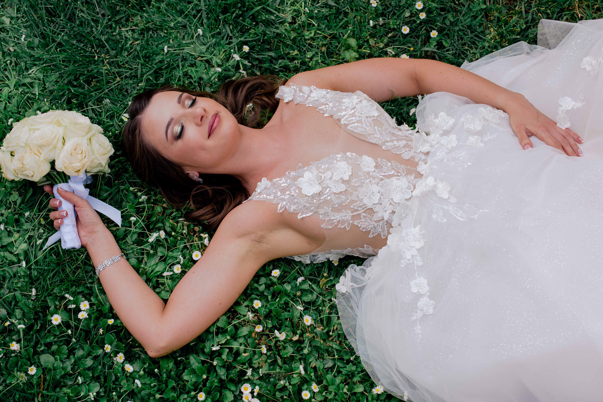 Panna młoda w białej sukni leży na trawie z bukietem białych róż, delikatny makijaż oczu z błyszczącym cieniem i bransoletka na ręce