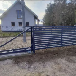 Nowoczesna, przesuwna brama wjazdowa w kolorze grafitowym, z poziomymi panelami, na tle nowego domu jednorodzinnego i siatki ogrodzeniowej. Brama częściowo otwarta.