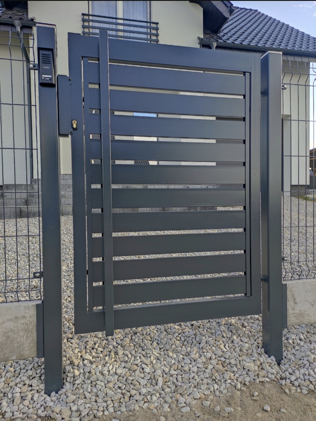 Nowoczesna, grafitowa brama wjazdowa z poziomymi szczeblami, zamontowana przy posesji z ogrodzeniem z siatki. Widoczny dom w tle i podłoże z białego kamienia.