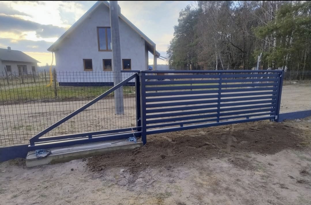 Nowoczesna, przesuwna brama wjazdowa w kolorze grafitowym, z poziomymi panelami, na tle nowego domu jednorodzinnego i siatki ogrodzeniowej. Brama częściowo otwarta.
