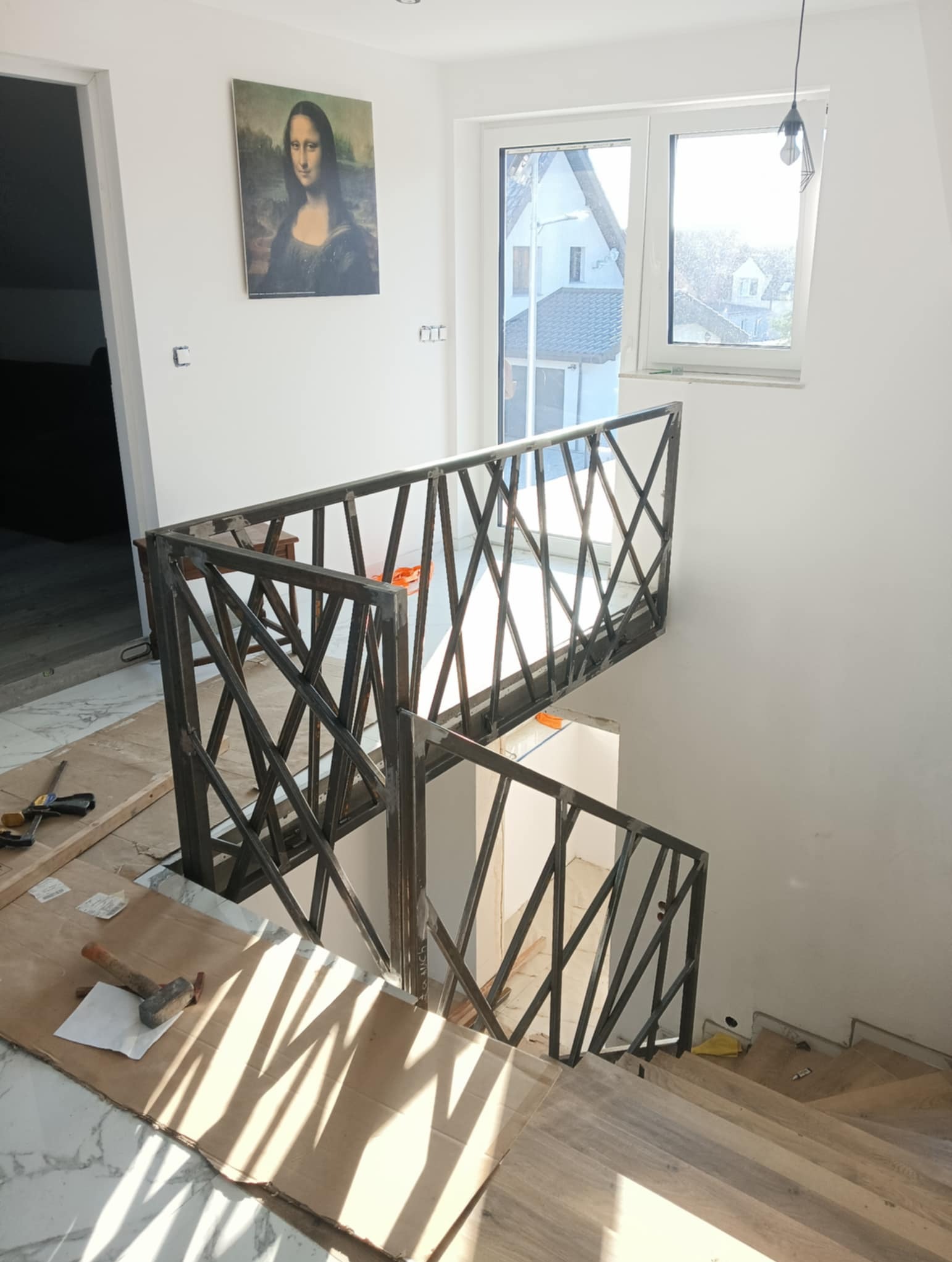 Metalowa balustrada schodowa o geometrycznym wzorze w surowym wnętrzu z obrazem Mony Lisy. Widoczne narzędzia sugerują trwający montaż.