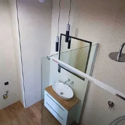 Nowoczesna łazienka z drewnianą podłogą, owalną umywalką nablatową na białej szafce z drewnianym blatem, kabiną prysznicową ze szklaną ścianką i dwoma wiszącymi lampami z czarnymi elementami.