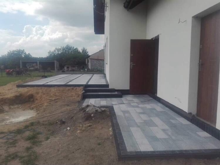 Układanie kostki brukowej w odcieniach szarości i czerni na podjeździe domu, widoczne schody z kostki, teren w trakcie prac ziemnych.