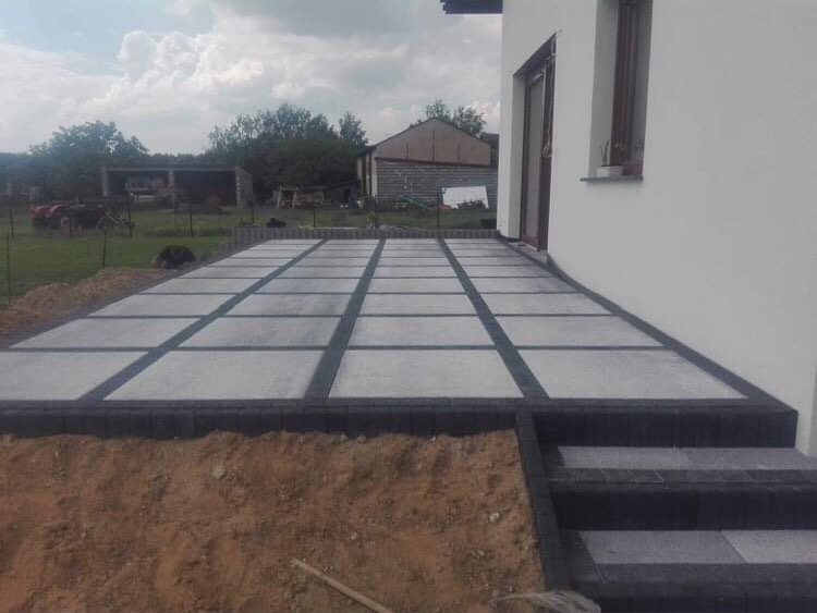 Ukończony taras z jasnoszarych płyt betonowych oddzielonych ciemnoszarymi kostkami, prowadzący do wejścia do białego budynku, z widocznymi dwoma stopniami schodów.