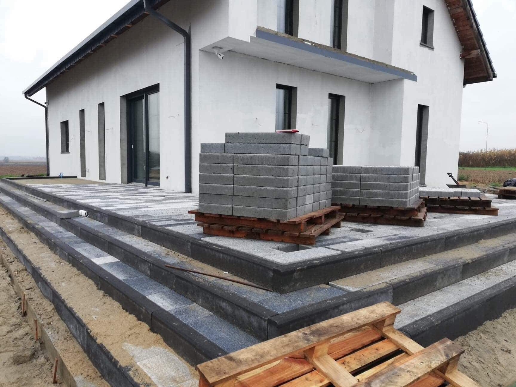 Schody wejściowe do domu wyłożone kostką brukową w dwóch odcieniach szarości, z paletami kostki czekającymi na ułożenie, widok z boku na dom.