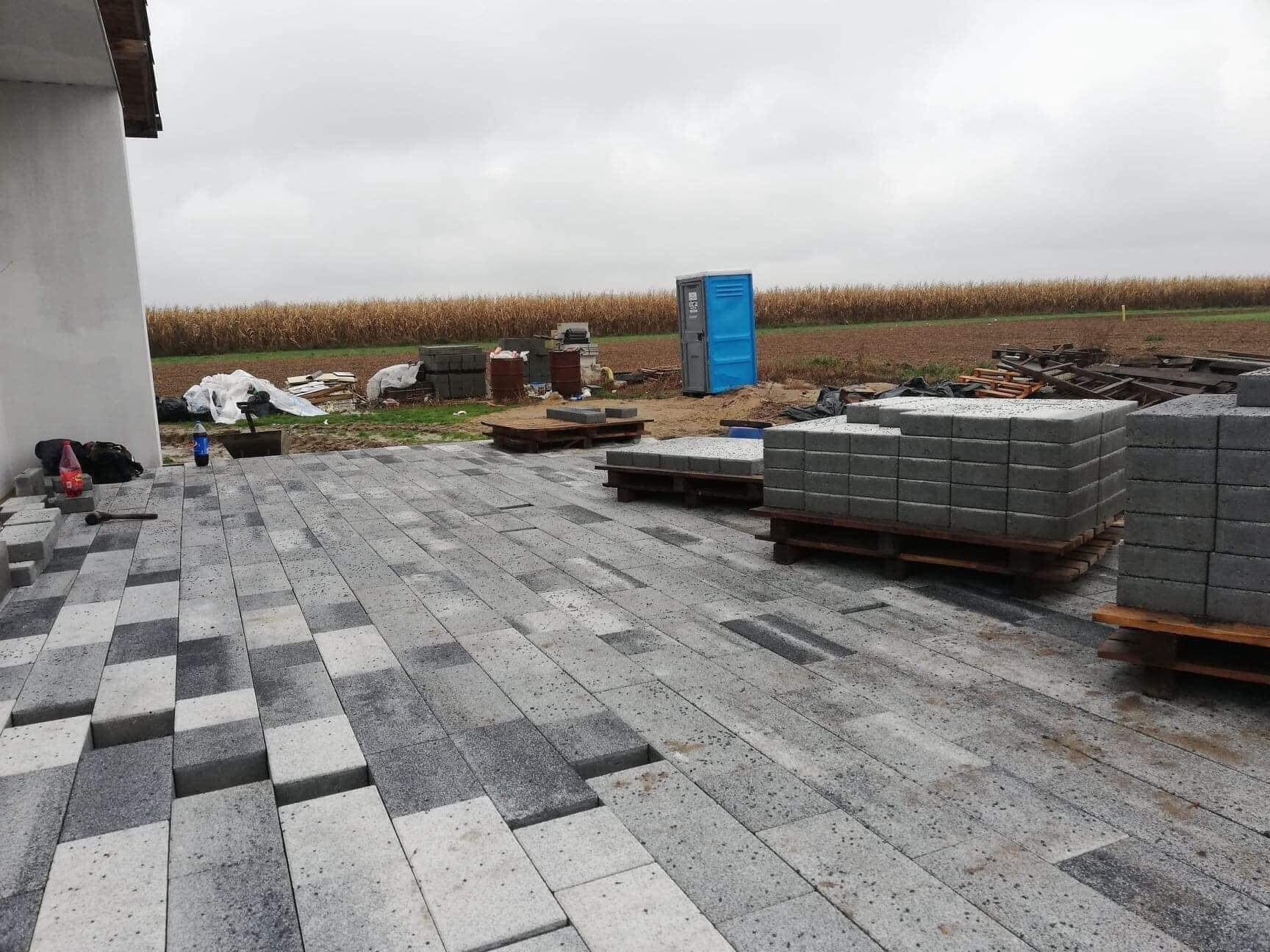 Niedokończona nawierzchnia z kostki brukowej w szaro-białej kolorystyce, z widocznymi paletami z kostką w tle, na tle pola kukurydzy i przenośnej toalety.