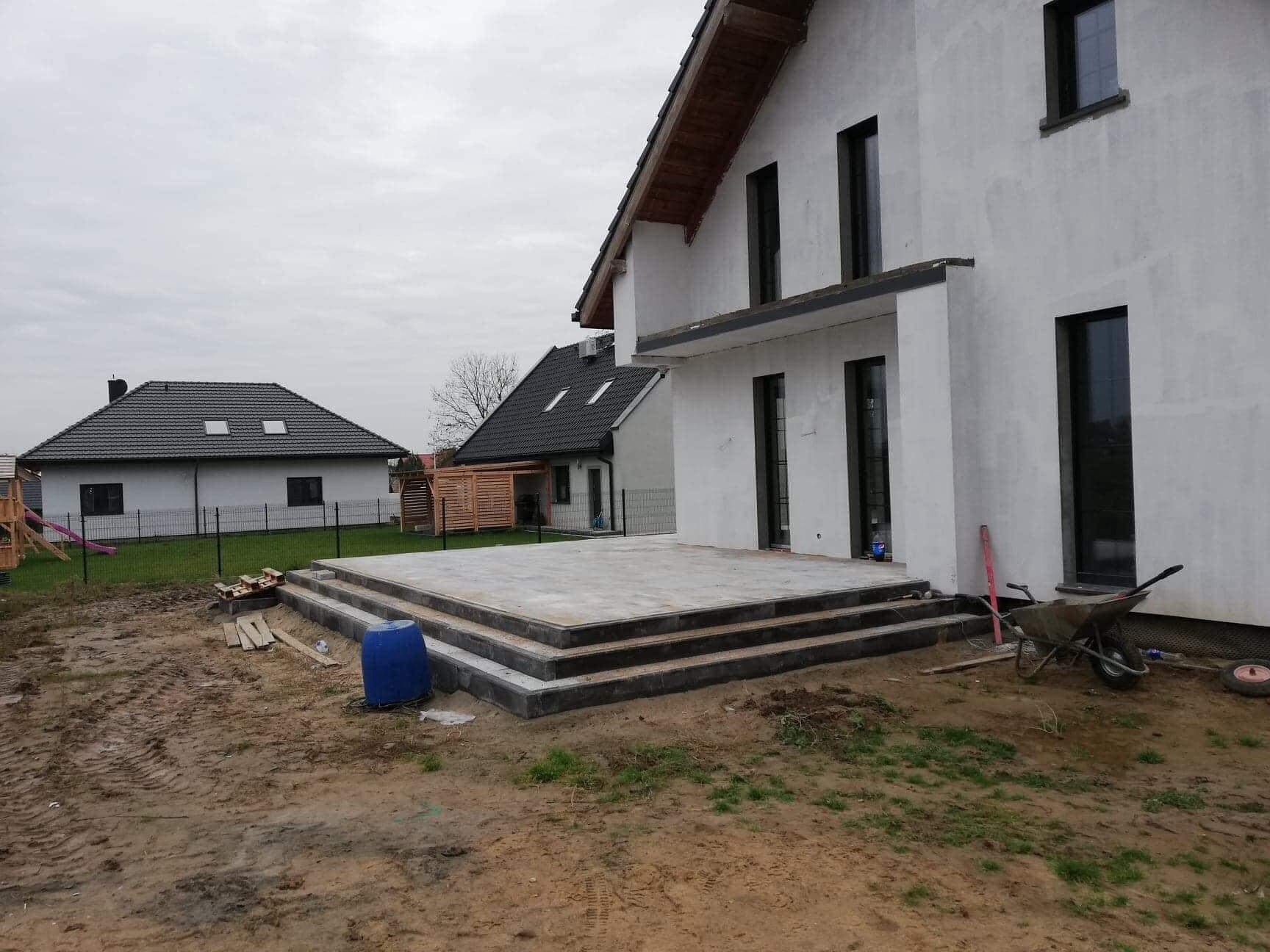 Trzy stopniowe schody z szarej kostki prowadzące do domu w trakcie budowy, widoczny niebieski pojemnik i taczka z narzędziami na gliniastym podłożu.