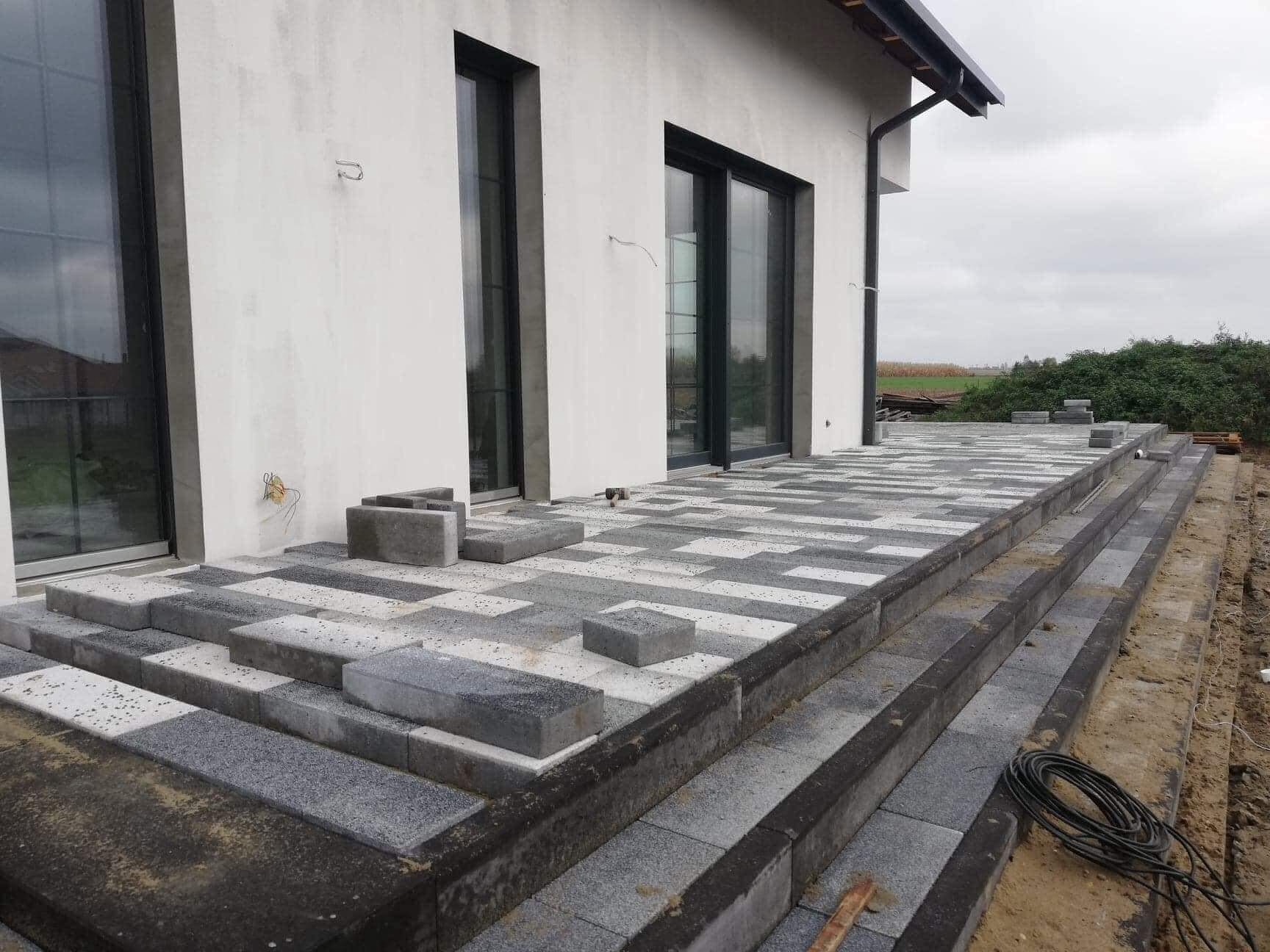W trakcie tworzenia tarasu z kostki brukowej w geometryczny wzór, widoczne niedokończone schody i przygotowane elementy kostki przy nowoczesnym domu z dużymi oknami.