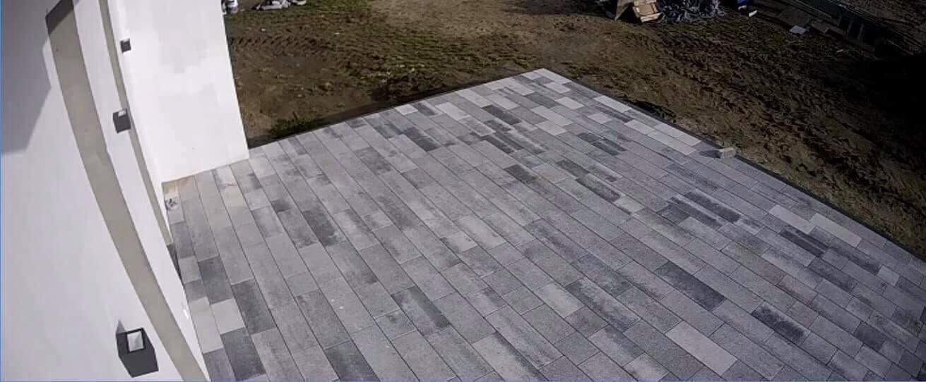Ułożona szara kostka brukowa wzdłuż ściany budynku, widok z góry, nierówna powierzchnia terenu w tle.
