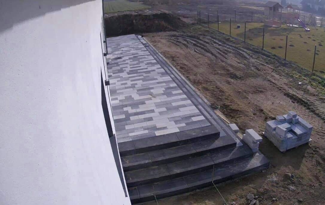 Nowoczesna nawierzchnia z jasnoszarej i ciemnoszarej kostki w prostokątnym wzorze, prowadząca do wejścia budynku z trzema schodami, obok sterta nieużytej kostki, widok z góry.