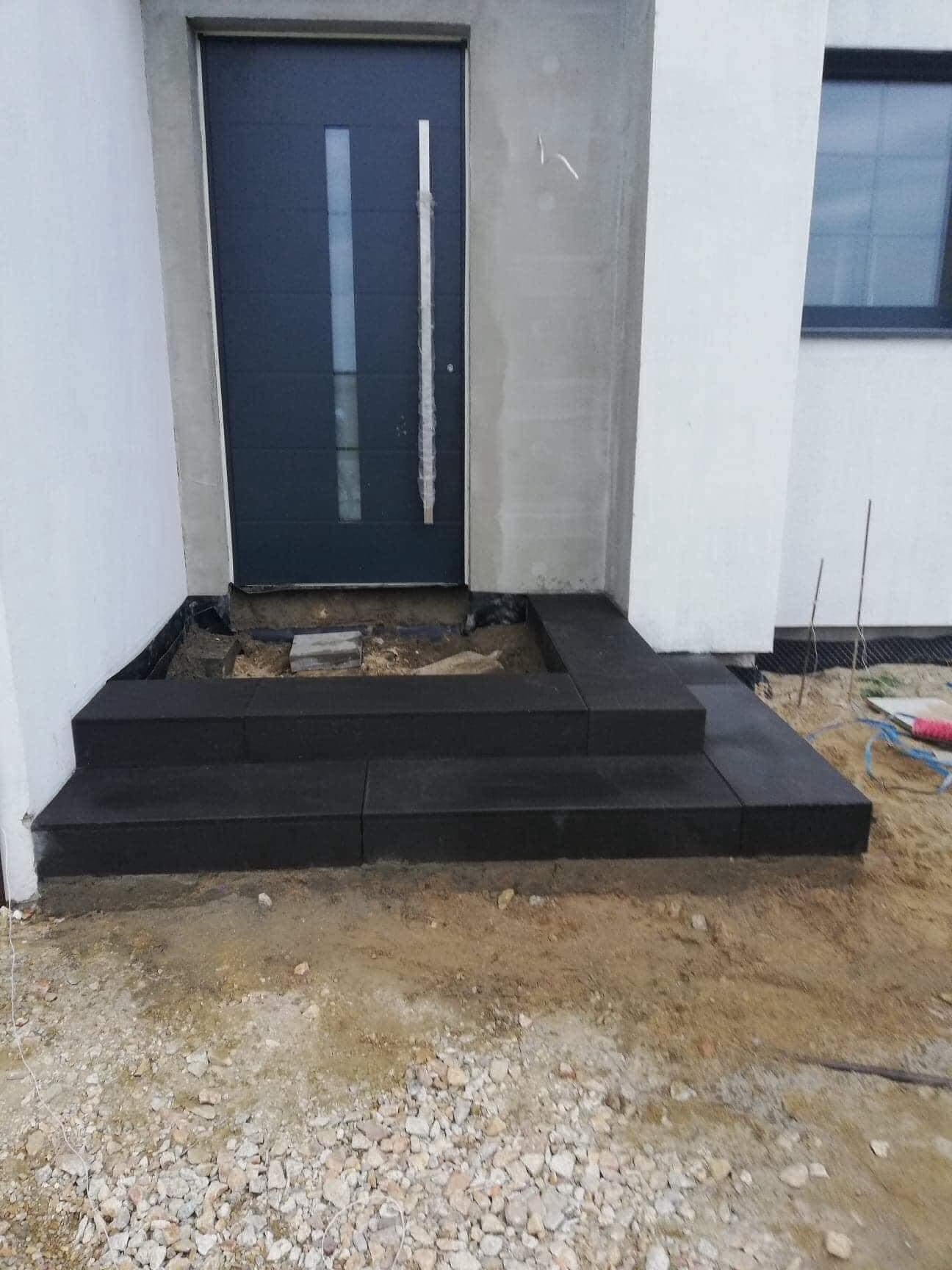 Nowe, ciemne, kamienne schody wejściowe prowadzące do nowoczesnego domu z szarą stolarką drzwiową, widoczne niedokończone prace ziemne.