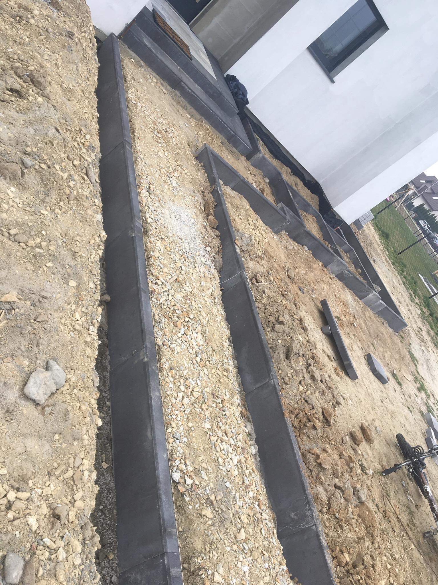 Przygotowanie podłoża pod ścieżkę z obrzeżami, widoczne ułożone krawężniki i podsypka piaskowa, w tle fragment budynku z oknem.