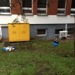 Dwa klimatyzatory typu split zamontowane na zewnątrz budynku przychodni, obok żółtej skrzynki gazowej i narzędzi serwisowych.