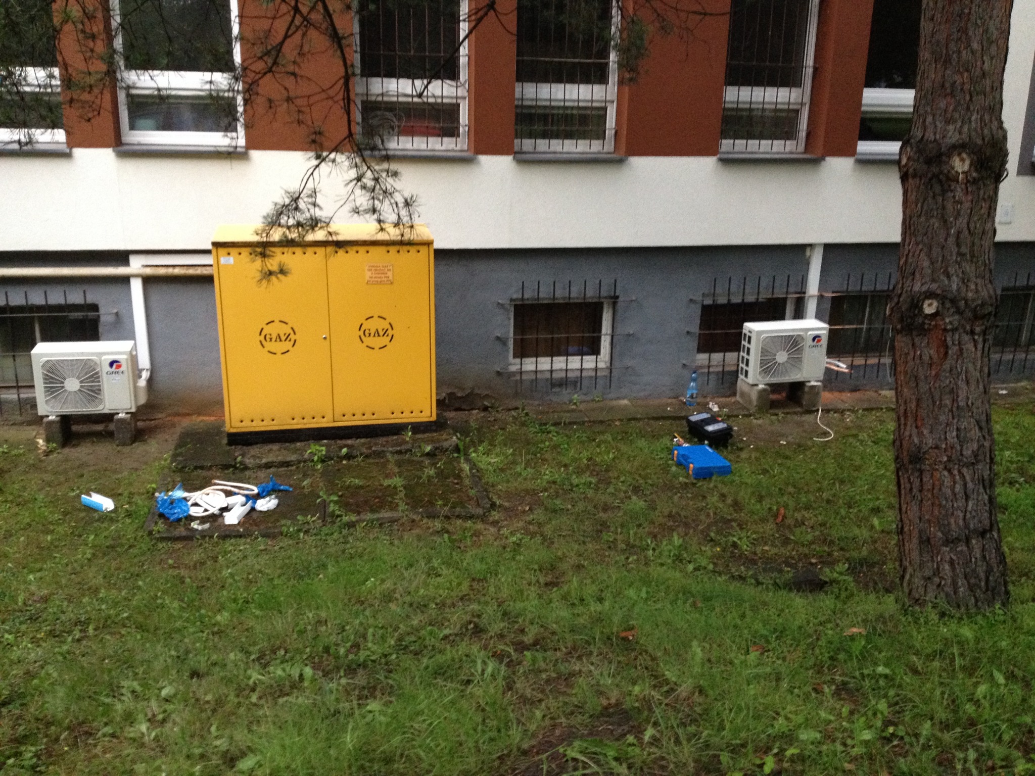 Dwa klimatyzatory typu split zamontowane na zewnątrz budynku przychodni, obok żółtej skrzynki gazowej i narzędzi serwisowych.