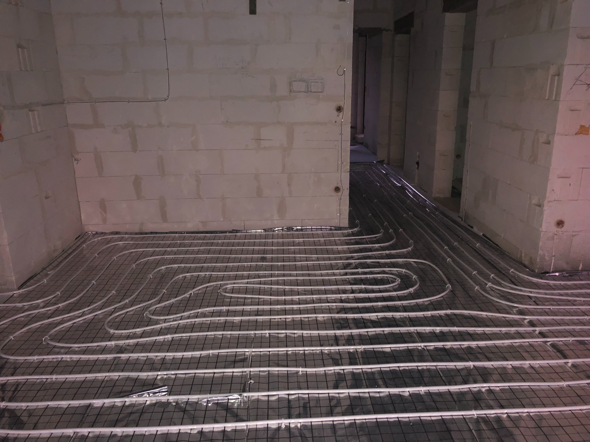 Montaż ogrzewania podłogowego w surowym stanie, rury grzewcze ułożone na czarnej siatce w pomieszczeniu z białymi ścianami z betonu komórkowego.