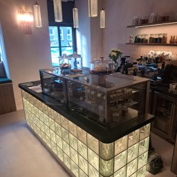 PE - Smart Home - Nowoczesny wystrój kawiarni: lada z luksferami, szklane witryny z ciastami, designerskie lampy wiszące i subtelne oświetlenie półek. Pastelowe ściany i jasna podłoga.