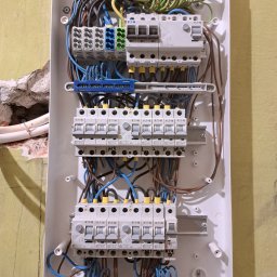 PE - Smart Home - Rozdzielnica elektryczna w białej obudowie, z widocznymi bezpiecznikami, przewodami w kolorach: niebieskim, brązowym, czarnym i żółto-zielonym. Ściana w tle jest żółta.