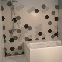 Nowoczesna łazienka z heksagonalnymi płytkami w odcieniach szarości i bieli na ścianach i podłodze, biała wanna zabezpieczona folią ochronną oraz brodzik z odpływem i chromowana bateria prysznicowa.