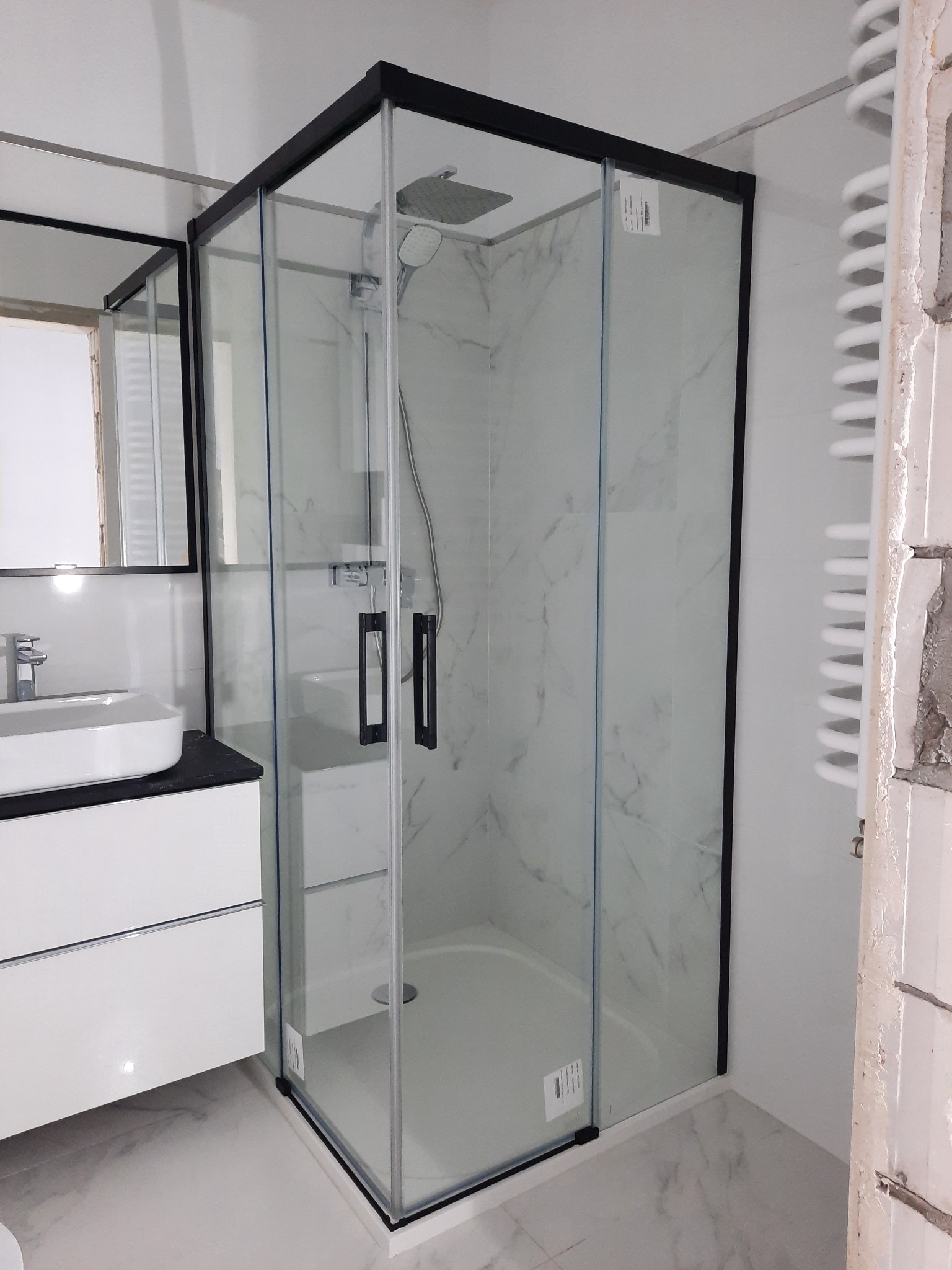 Nowoczesna kabina prysznicowa z czarnymi profilami i marmurowym wzorem wewnątrz, zamontowana w łazience w Warszawie, obok umywalki z szafką i grzejnika drabinkowego z widocznymi śladami prac...