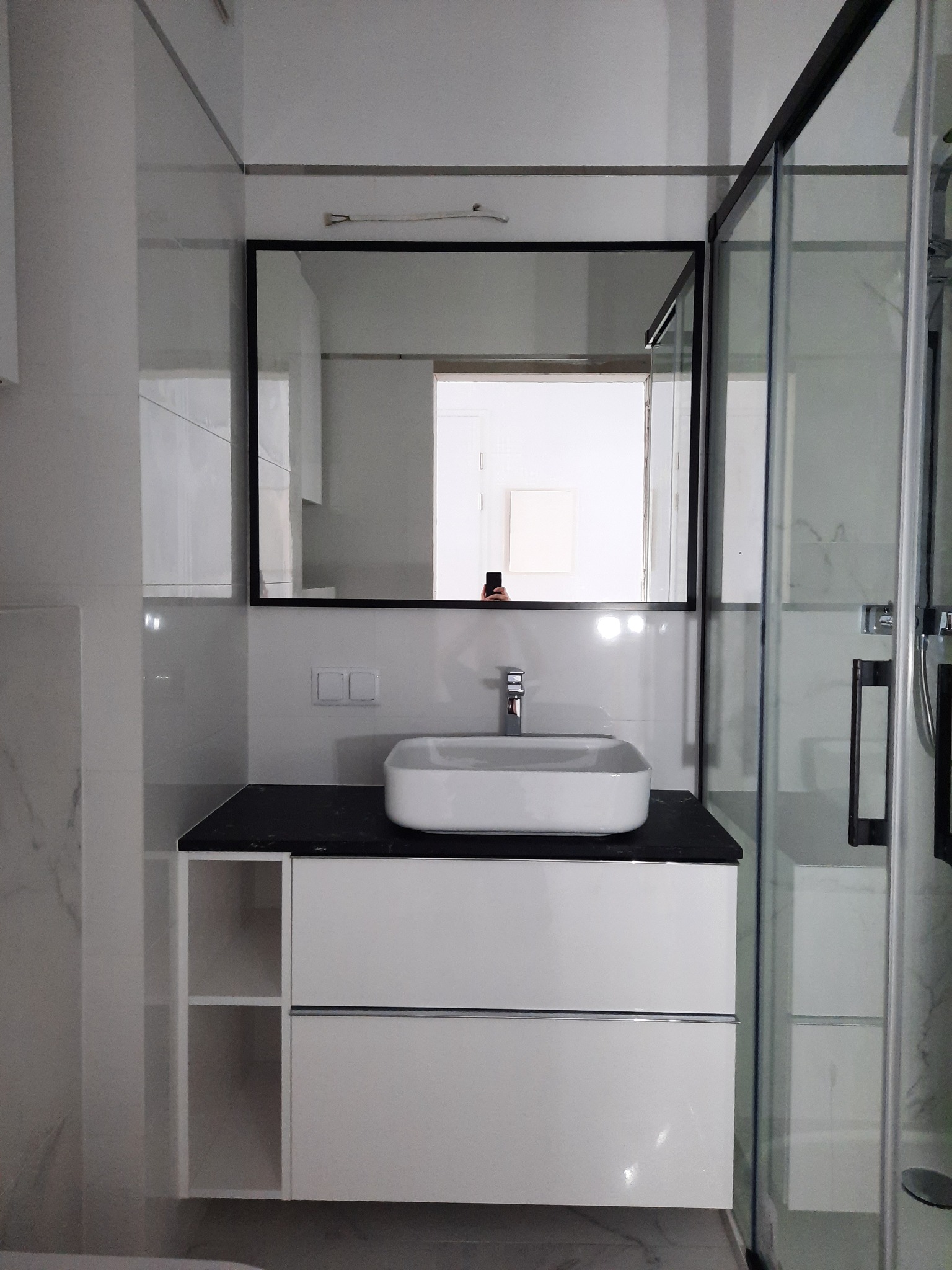 Nowoczesna łazienka z białą szafką pod umywalkę z czarnym blatem, prostokątną umywalką nablatową i czarną ramą lustra, kabina prysznicowa ze szklanymi drzwiami.
